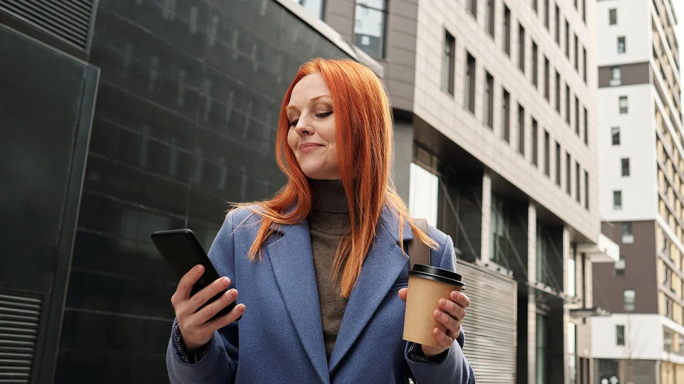 Person using a smartphone while holding a coffee cup outside a city building.